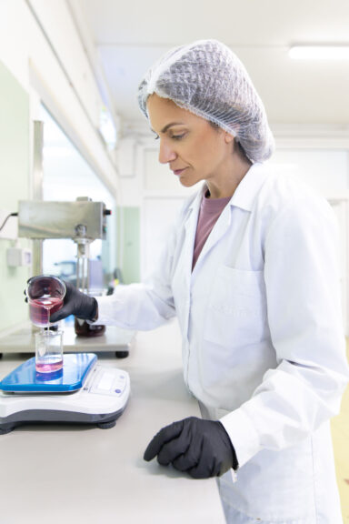 Tecnica di laboratorio Cosmetu che versa una soluzione rosa in un cilindro graduato su bilancia, durante la formulazione di prodotti cosmetici
